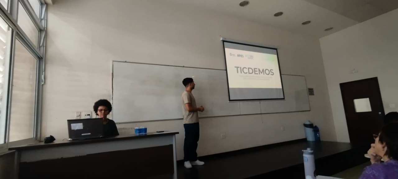 Sala de aula com o Claudio, homem branco de camisa verde, calça jeans azul e tenis branco, apresentando um trabalho com o slide escrito "TICDEMOS" e Ana Júlia, mulher negra, no computador